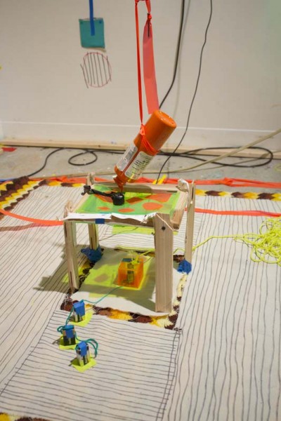 A small chair made of wood with an orange bottle suspended in the air with a red ribbon, a green sheet on the chair with orange spots, a battery pack under the chair wired with a blue and green line put in the middle of the floor surrounded with papers with black lines drawings