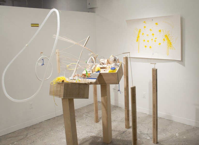Installation view of many objects on wooden stand, made of organic blue, yellow or white material, a hose hun gin the air with an invisible line, and a painting with yellow splashes on the wall