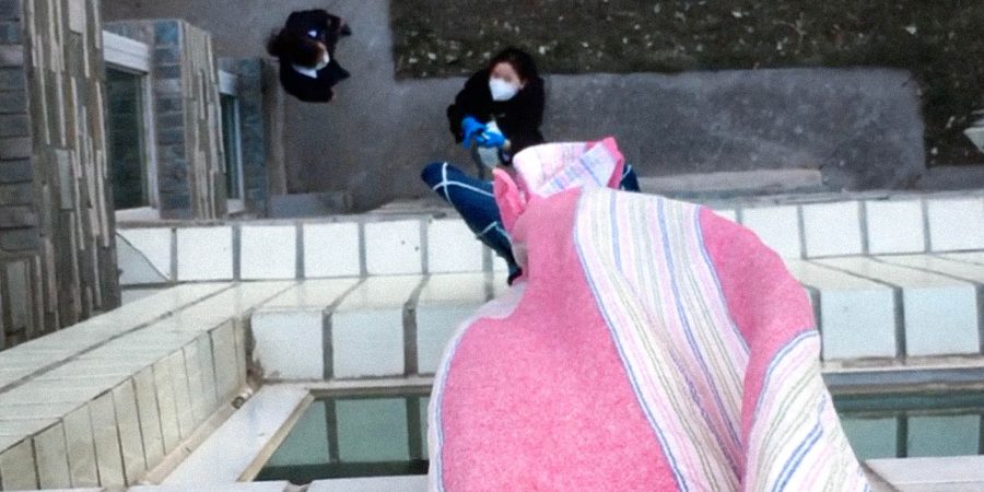 Hanni Huang pulling supplies up to a window in a quarantine hotel. The photograph was taken from the vantage point of the person looking down at the ground from the window. A pink and white blanket is hanging out of the window and someone is standing on the ground looking up at it.