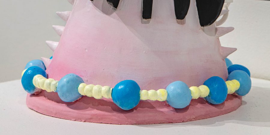 The bottom portion of a sculpture consisting of a pink cone with a blue and yellow beaded string wrapped around the base. Spikes line the sides of the cone.
