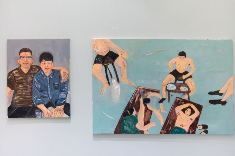 Two paintings representing people are installed on a white wall. The one on the left side represent two friends sitting on a bench, with a blueish-grey background, and the other one represents a few people sitting on chairs and two laypersons on maroon blankets on the ground