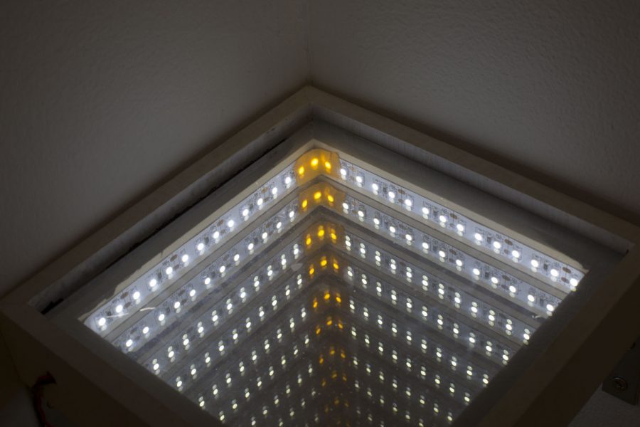 A square frame that is showing white circular lights with three yellow lights in the center, the sculpture is installed with a mirror so it looks like an infinity of lights when you peer through it