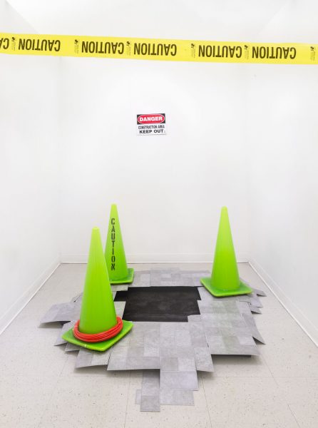 On the wall there is yellow caution tape installed, below is a white sign that says "Danger" in red and "Keep out" in black text, in front of this on the floor are a bunch of gray tiles with a black hole in the center, there are three neon green traffic cones around it, one says caution printed vertically on the cone, another cone has a neon orange rope on the base of it
