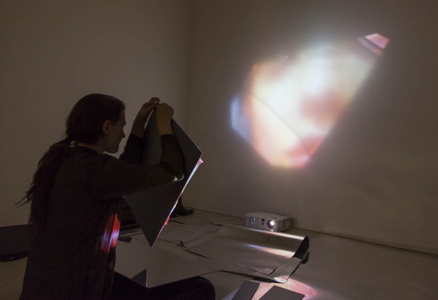 A student sitting on the ground working with a reflective foil that reflects an image on the wall from a video projector sitting on the floor.