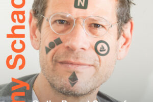 A photograph of artist, writer and critic, Kenny Schachter. The photograph is a headshot of Kenny with digital tattoo-like images and text across his face. The text on his forehead says the words, "Nifty Gateway".