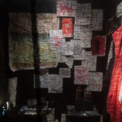 An art installation by Seung Won Lee titled "Drawing in hospital." The photograph was taken through a sheer black fabric and shows drawings and papers tacked to a wall with clothing hanging from hooks in a dark room lit by a single light bulb.
