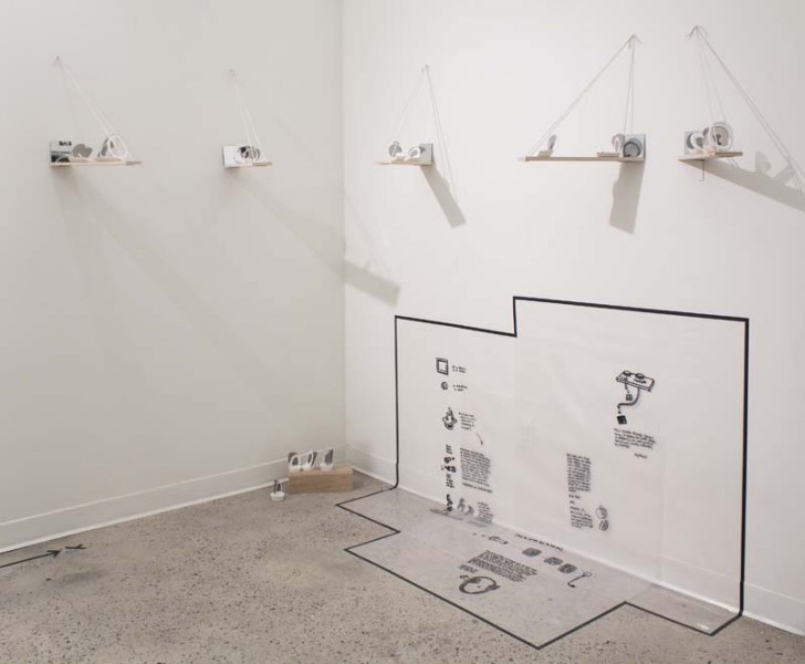 Installation view of mechanical parts drawn on the wall with a black rectangular shape as a frame, and eight wood pieces with black graphics standing on the floor.