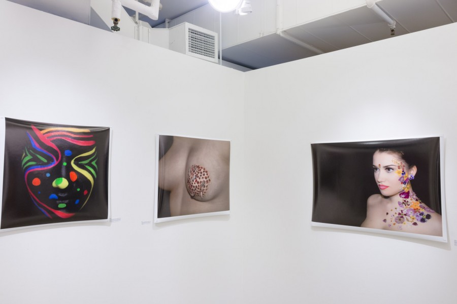 Three prints hung on the wall of a face painted in black and red, green, blue lines and dots on it, a side view of a woman's breast with the nipple covered in fabric with small organic shaped objects on it, and a portrait of a woman with many shapes in vivid colors beside her ear going down the neck and shoulder.