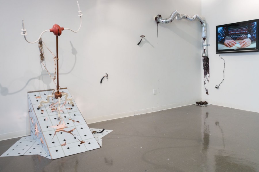 Installation view of a sculpture made with a white base with square outlines, a long stick fixed on the inclined part of the sculpture has two lights fixtured at its top. On the right side is a duct coming from the wall with a brown material leaking out of it, and next to it on the far right side is a TV with an image of a person holding an object with both hands on a table