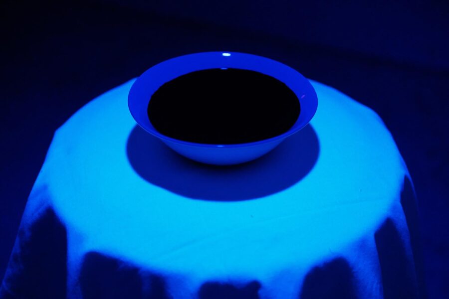 A sculpture by Steve Chen consisting of a ceramic bowl filled with ink on a small round table illuminated with black light.