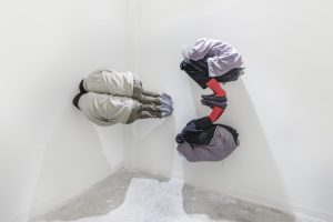 Sculpture of two people with their knees bent and face in one of their arms, there are plastic bags over their heads, and they are installed against the white wall connected at the feet so one is right side up and the other is upside down like a reflection of the first one, to the left there is a sculpture of two people right next to each other in the same pose, but stuck on the side and facing the gallery wall so you can't see their front