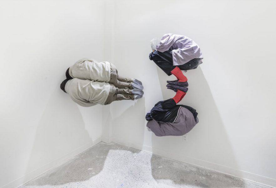Sculpture of two people with their knees bent and face in one of their arms, there are plastic bags over their heads, and they are installed against the white wall connected at the feet so one is right side up and the other is upside down like a reflection of the first one, to the left there is a sculpture of two people right next to each other in the same pose, but stuck on the side and facing the gallery wall so you can't see their front