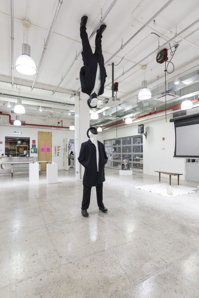Sculpture of a person standing up and looking up, wearing black blazer, white shirt, black pants and black shoes, the face is covered in black with a black hat, the piece is looking up and there is a second piece that is similar except the face is not covered in black and it is standing upside down on the ceiling right above the first figure sculpture