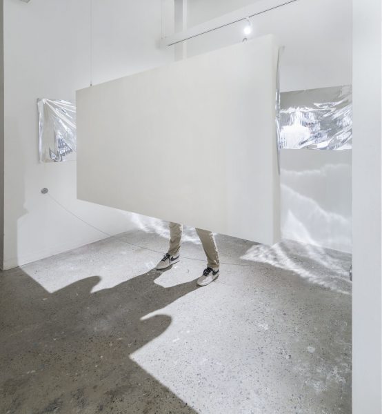 Installation there is a white rectangle that is haging with someone standing behind it wearing white pants and white and black sneakers, it looks like there are a few projections behind the piece of clouds or mountains that are also white and gray