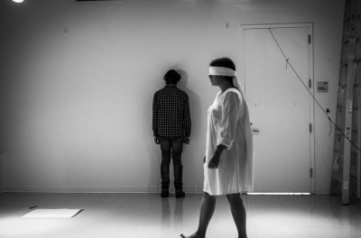 A shot representing a person standing in front of a wall facing the wall, a closed-door on their right side, and a person with eyes covered wearing a hospital shirt crossing the room from right to the left, facing to the left part of the room