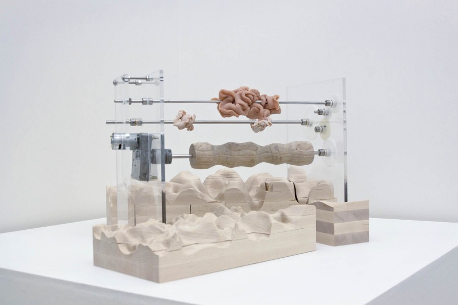 A device with a wooden base and a few metal rods holds three organic shaped orange and nude color sculptures
