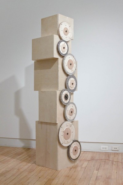 View of a wood sculpture made of cubes, and the cubes have mixed-sized gears attached on the front side of the sculpture.