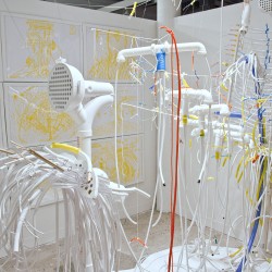 Installation view of white plumbing lines hung with invisible fishing line, mounted on the wall, and in the background is a grid of paintings made with yellow on white background in white frames.