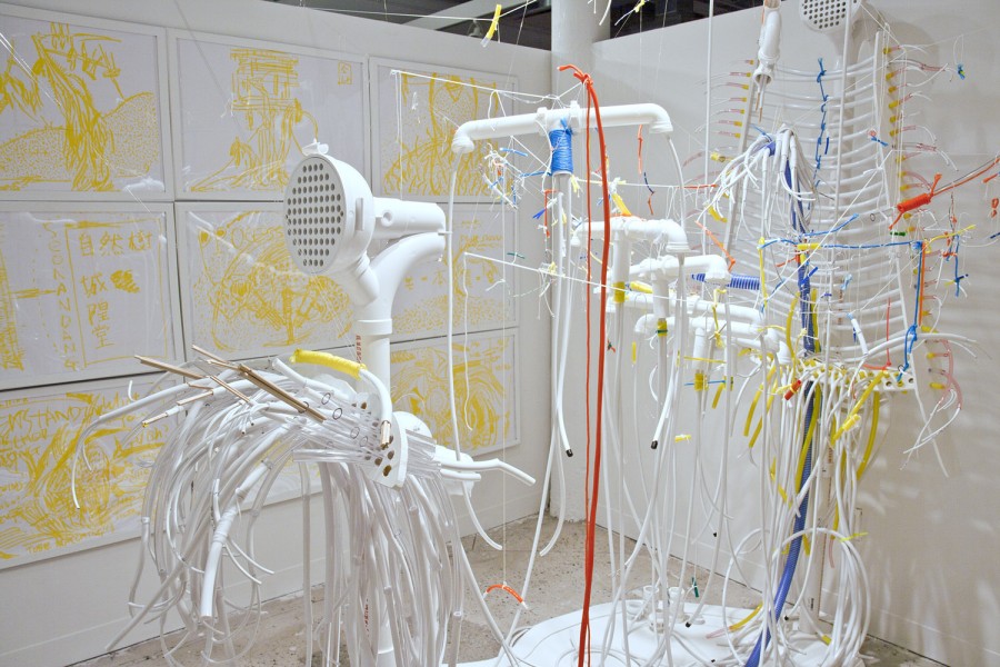 Installation view of white plumbing lines hung with invisible fishing line, mounted on the wall, and in the background is a grid of paintings made with yellow on white background in white frames.