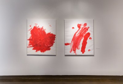 Two paintings by Amy Destefano hang on a wall. The paintings feature a solid white background with red paint splashed across the surface in an arbitrarily-looking manner.