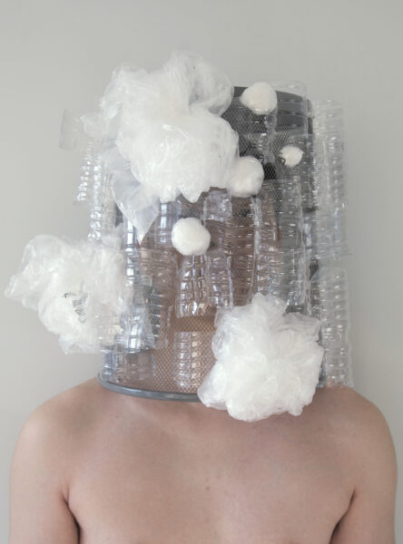 A photograph of a shirtless person wearing an inverted wire-mesh dust bin covered in bunches and balls of cellophane and bubblewrap.