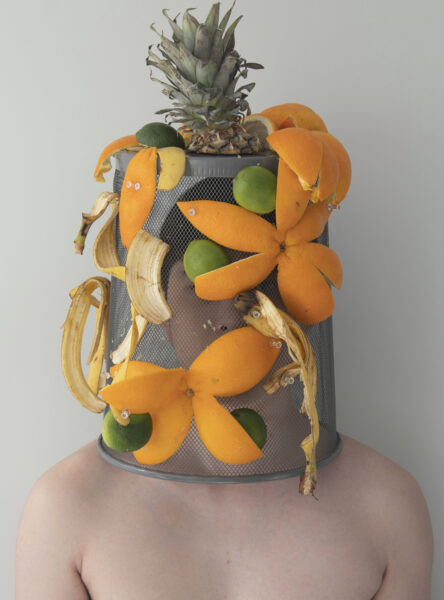 A photograph of a shirtless person wearing an inverted wire-mesh dust bin covered with orange, lime, and banana peels and topped with the leaves of a pineapple.