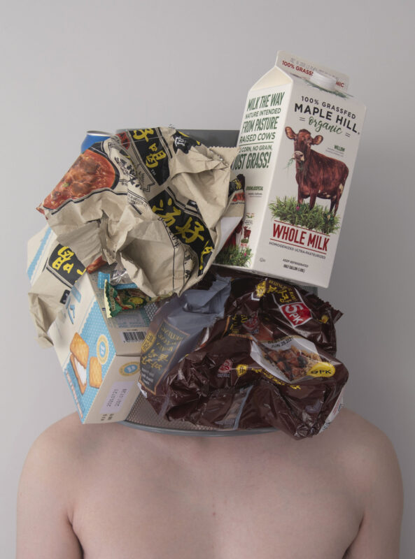 Color photograph of unknown male figure with metal trash bin over his head. There are various detritus covering the waste bin which consists of a milk container, cracker box, and crumpled snack baggies.
