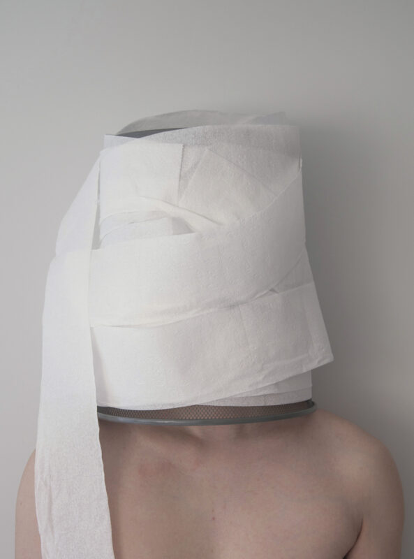 Color photograph of a white man's torso whose head is covered with a metal trash bin that has toilet paper wrapped around the exterior which hides the identity of the figure.