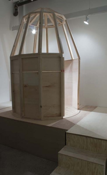 Installation view of a sculpture made of wood, wood boards, and wood staples, mounted in an angular shape with a round bottom, with the form of watchtower with the roof uncovered