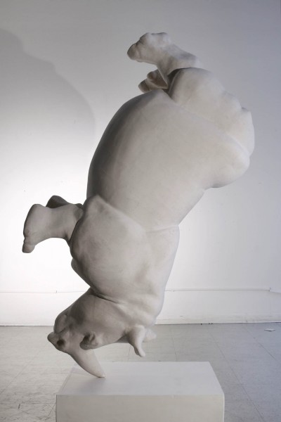 A different angle view of a rhino sculpture made of a white material which is positioned stick on its horn to a white plate on the ground
