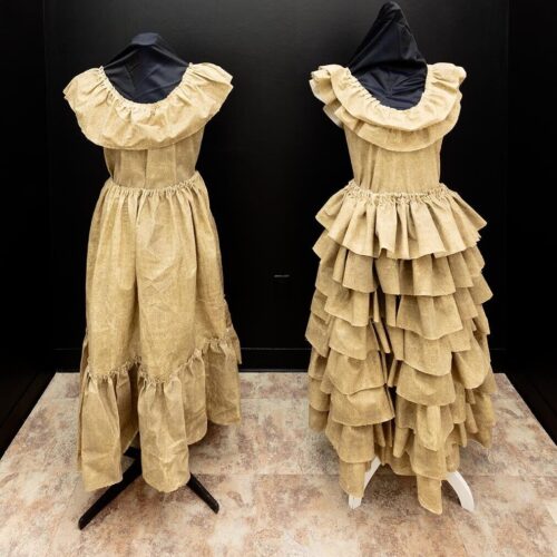 A pair of manikins wearing brown dresses in a room with black walls. The dresses are made from unbleached muslin fabric, sand, mold builder, thread, chicken wire.