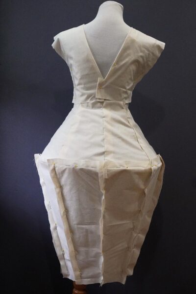 An off-white dress on a manikin viewed from the back. The dress is made from unbleached muslin fabric, sand, mold builder, thread, and chicken wire.