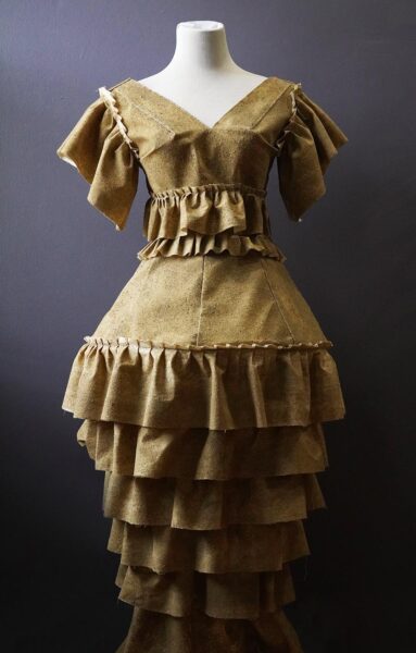 A manikin wearing a brown dress in front of a black wall. The dress is made from unbleached muslin fabric, sand, mold builder, thread, chicken wire.