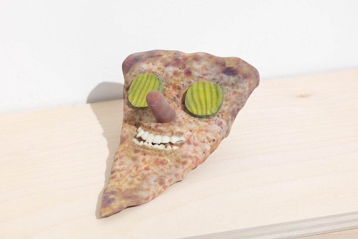 A 3D printed sculpture by Titus McBeath depicting a slice of pizza with a grinning face on it. The teeth appear lifelike but the eyes are sliced pickles and the nose is a hot dog.