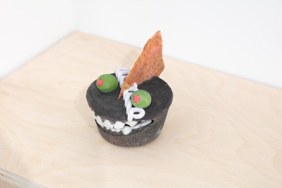 A 3D printed sculpture by Titus McBeath depicting a Hostess cake with olives and a tortilla chip on top. A mouth with teeth is embedded in the side of the cake so that the olives appear as eyes.