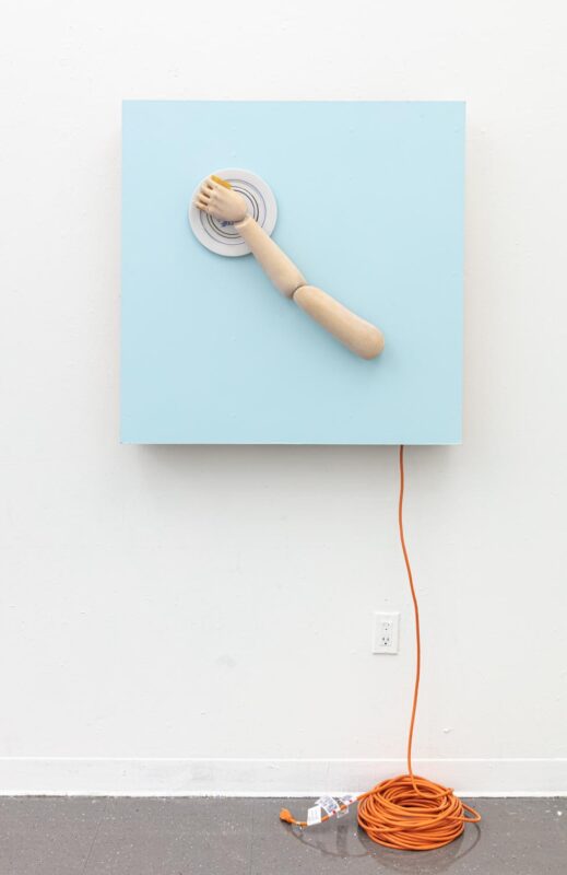 An artwork by Titus McBeath titled "Cain and Able (Able)" consisting of a light blue painting with a manikin arm holding a plate attached to it. A long orange extension cord is hanging from the back of the painting and is coiled up on the floor.