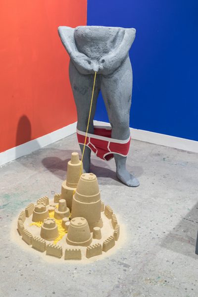 Installation view of artwork by Tony Seibert. A sculpture of the lower part of a grey male figure with yellow twine depicting urination and fabricated sandcastles.  