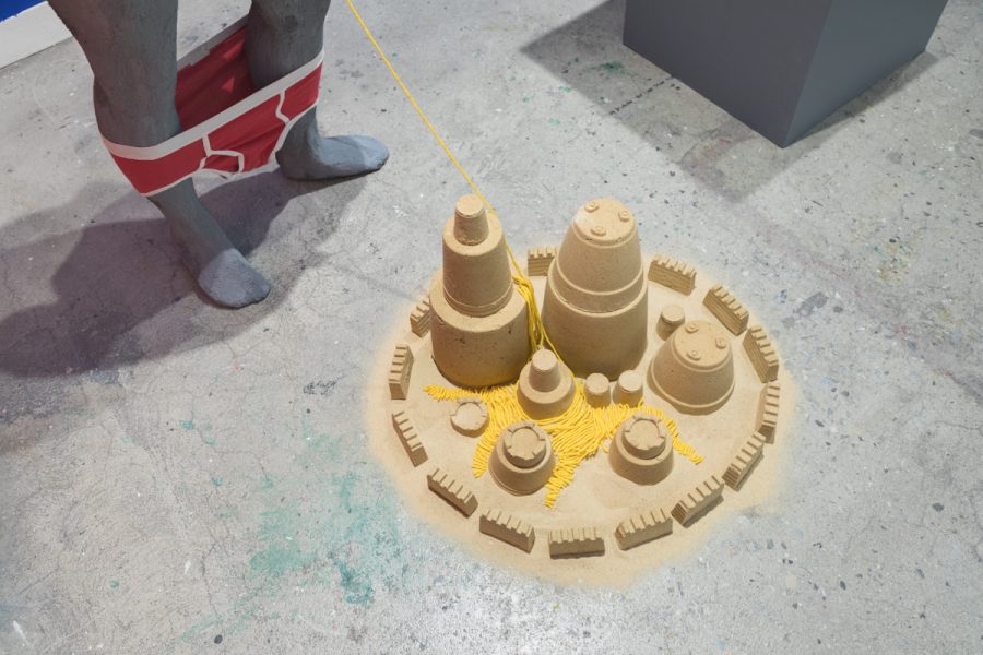 Installation view of artwork by Tony Seibert. Constructed sand castles placed on the floor with a grey figure with red male briefs lowered around the figures ankles.