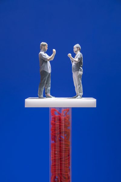 Sculptures by Tony Seibert. 3D printed figures facing each other placed on top of a fabricated hollow pedestal filled with red twine in front of a blue backdrop.