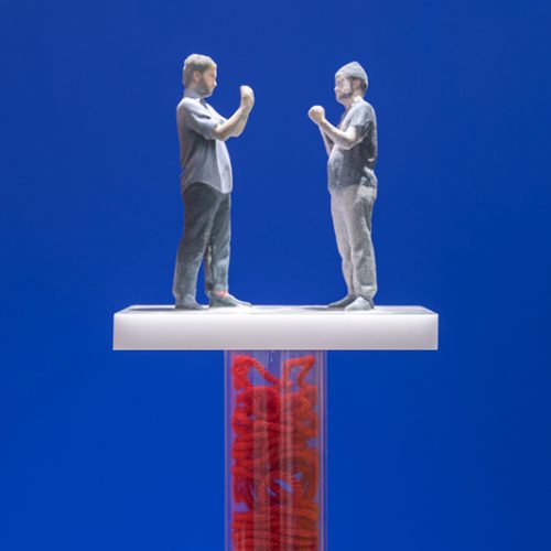 Sculptures by Tony Seibert. 3D printed figures facing each other placed on top of a fabricated hollow pedestal filled with red twine in front of a blue backdrop.