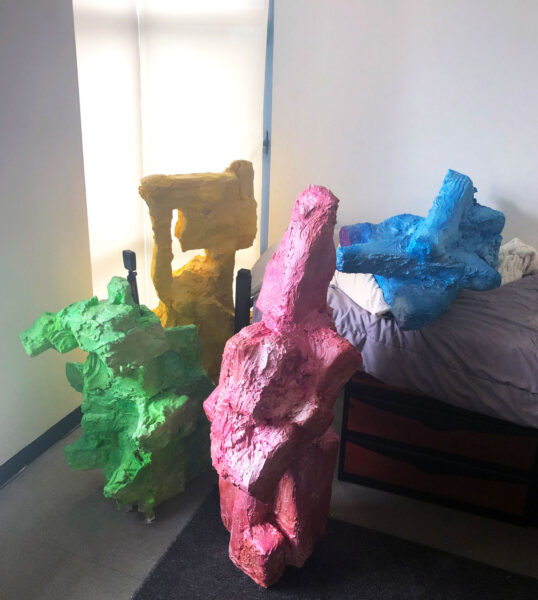 A set of four sculptures by Tyler Nicole Glenn displayed next to and on top of a bed. The sculptures are amorphous and each is a solid color (yellow, green, pink, or blue).