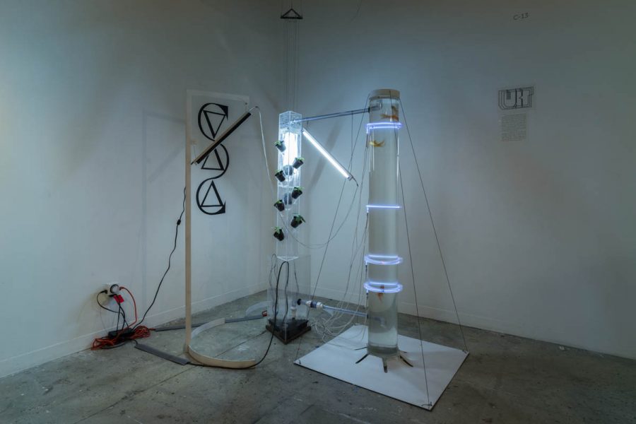Installation view of a tall tubular aquarium with goldfishes wrapped with white neon, beside is a triangular tower holding plants in black pots with soil and illuminated with white neon, and a woot structure with a metal rounded and angular part having a medium-long wood piece linked to the aquarium and the triangular structure