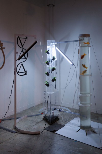 Installation vire of a sculpture made of a tall tube as an aquarium filled with water and four goldfishes. Beside is another tubular tower with a triangular aquarium at the base, filled with water. And on the left side is a wood structure with some triangles and a rounded-shaped metal, with a wood piece holding running water. In the back si a neon illuminating the sculptures
