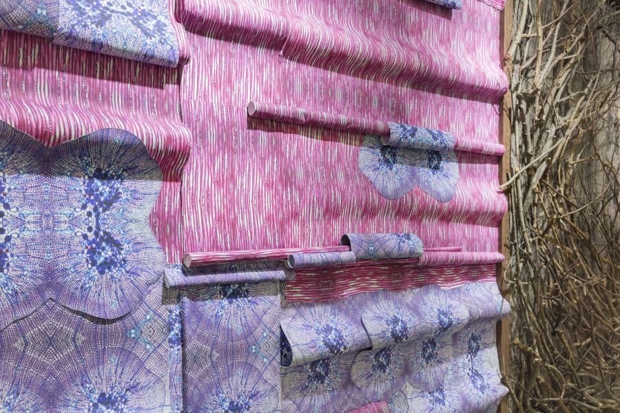 Close up of a wall piece depicting pink and purple wallpaper that is folded over on itself, the right there is a peek of a brown wall piece with branches on it
