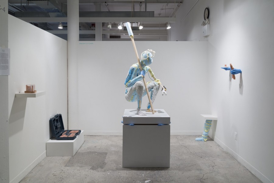 Installation view of an anthropomorphic sculpture in a crouch position with a long wooden stick in one hand