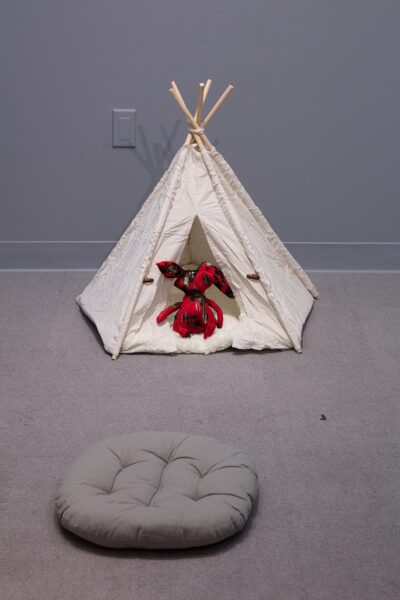 Installation by Lu with a tent sculpture and a red sculpture figure seated in the tent. Outside of the tent is a grey cushion.
