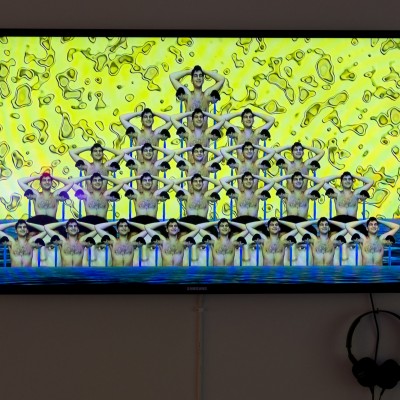 An array of many persons with their hands on their head organized in a triangle shape, on a yellow background, and at the bottom a portion of blue like-water color.