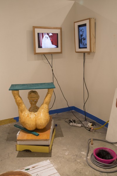 Installation view of two screens with images in a wooden frame on the wall and an orange sculpture of a human figure with feet in the air holding a green board.