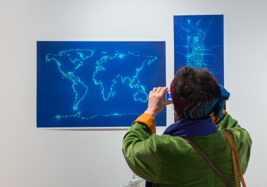 A person takes a photo of two drawings of a world map and a city map painted with fluorescent organic material on a blue background.