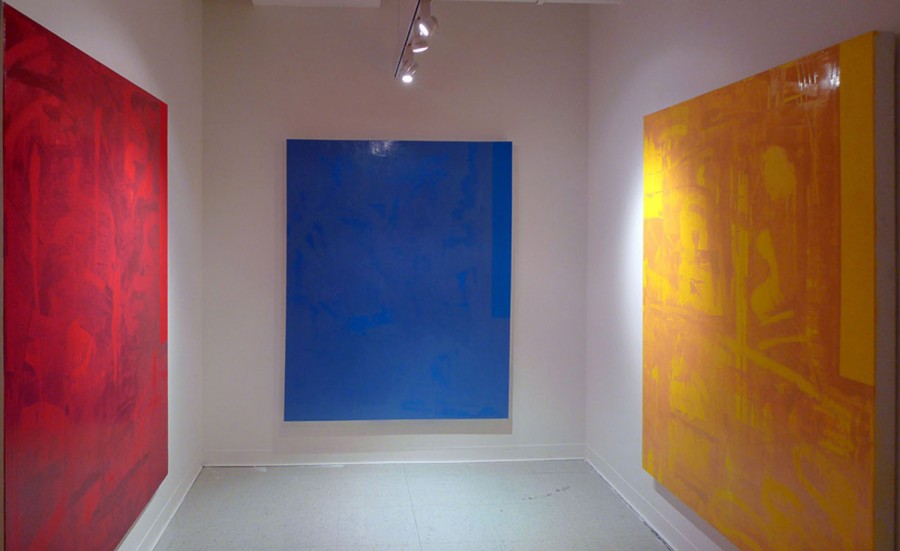 Exhibition view of three paintings, a red one on the left wall, a blue one in the front, and a yellow one on the right